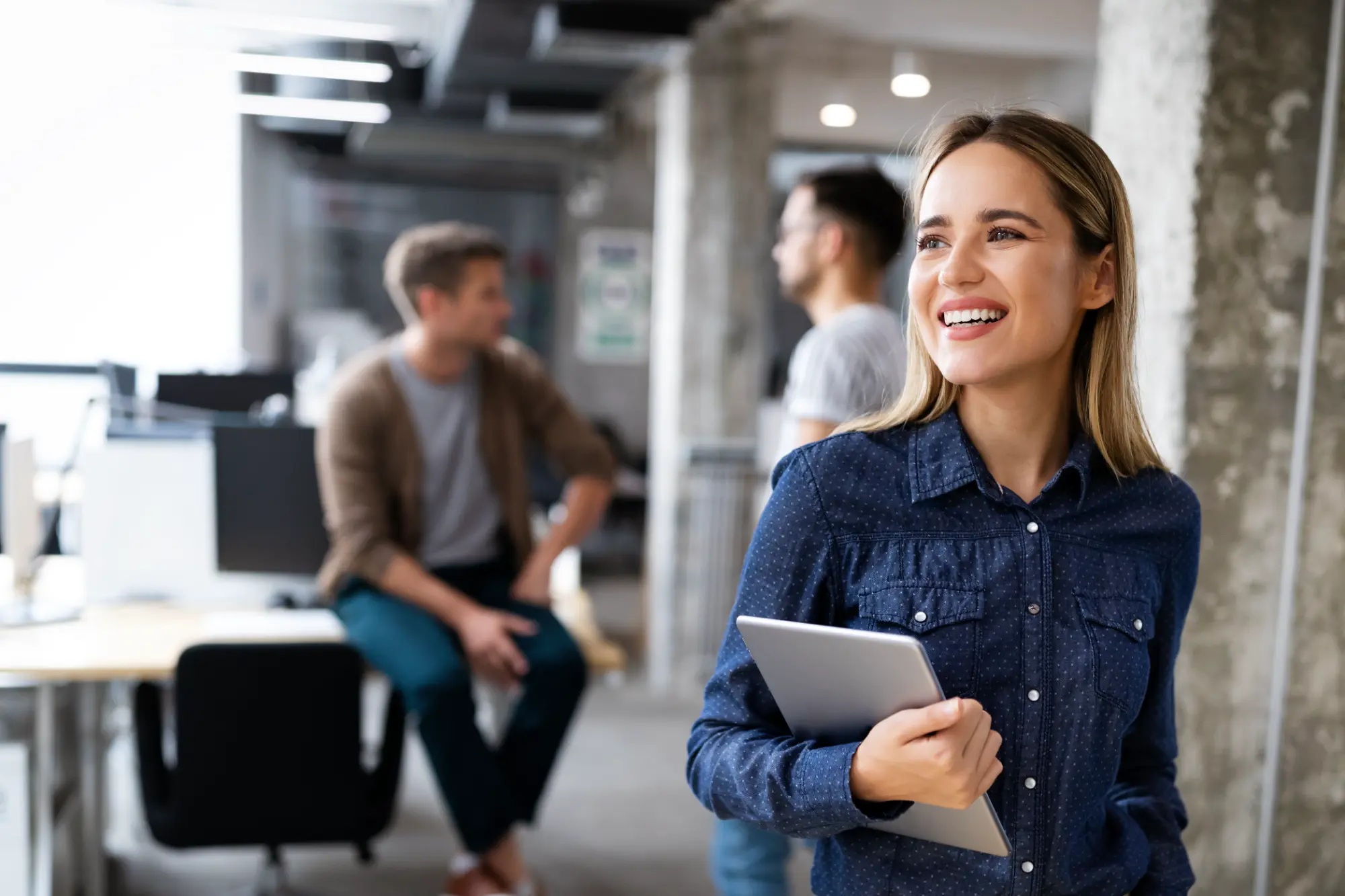 Young professional woman in office smiling and holding a tablet with colleagues blurred in background.