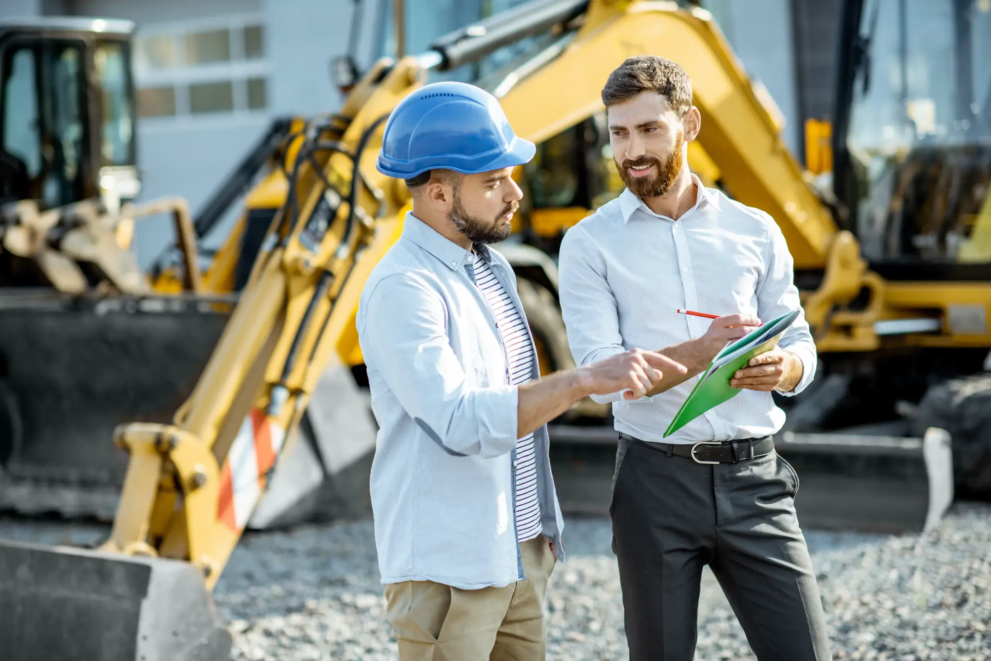 Construction workers and supervisor discussing something on a paper while standing in front of large construction vehicles.