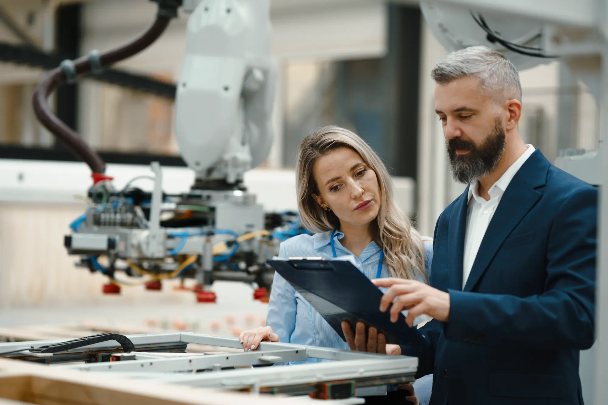 Middle-aged man and woman looking over clipboard around industrial machines.