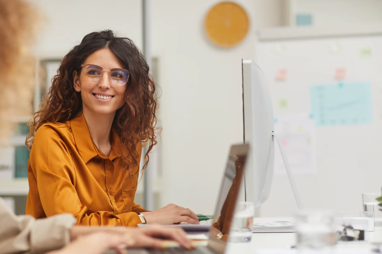 Young professional woman with glasses sitting at a computer smiling at a colleague.