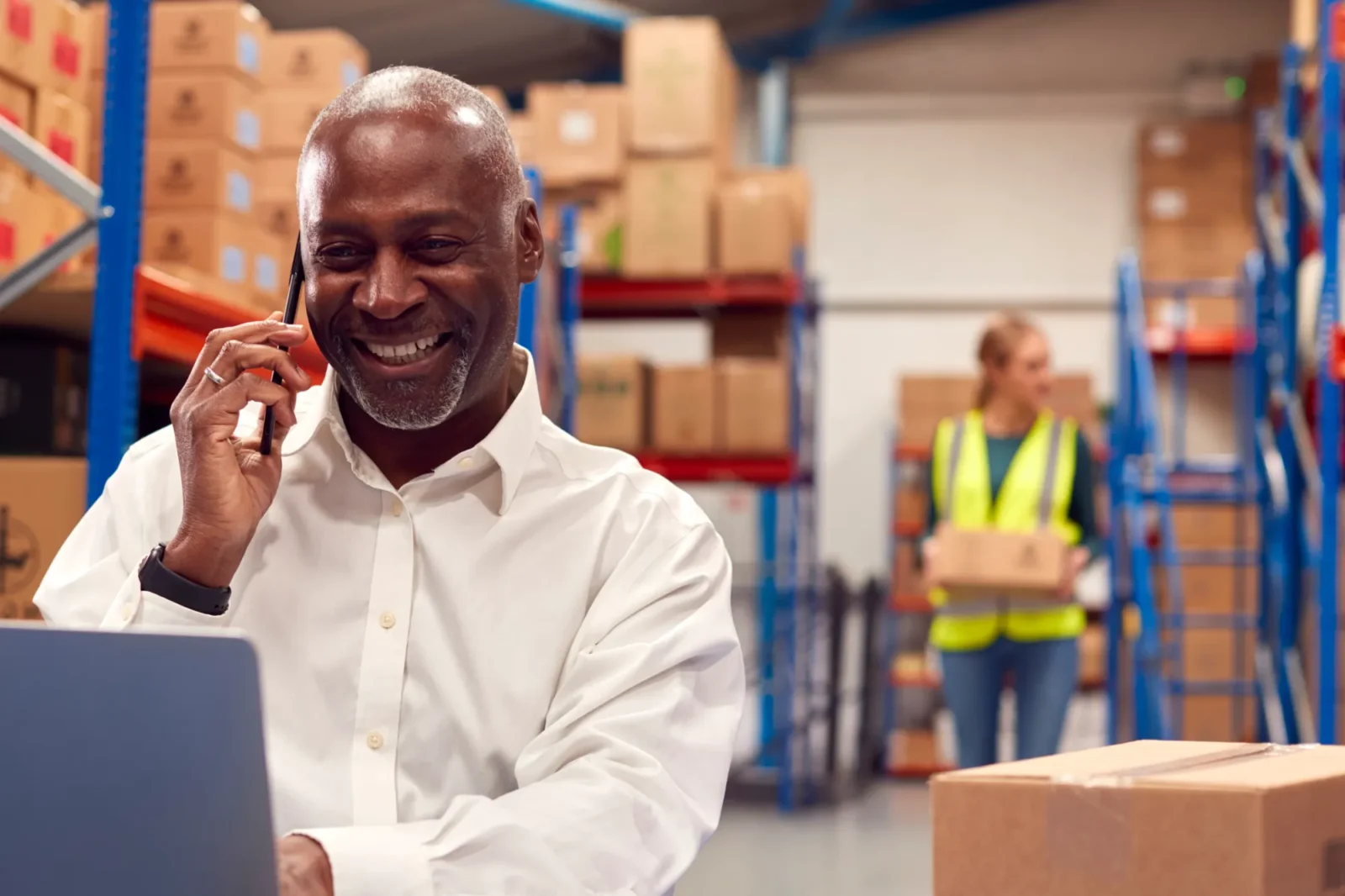 Older smiling man sitting at a desk inside a warehouse talking on phone and typing on a laptop.