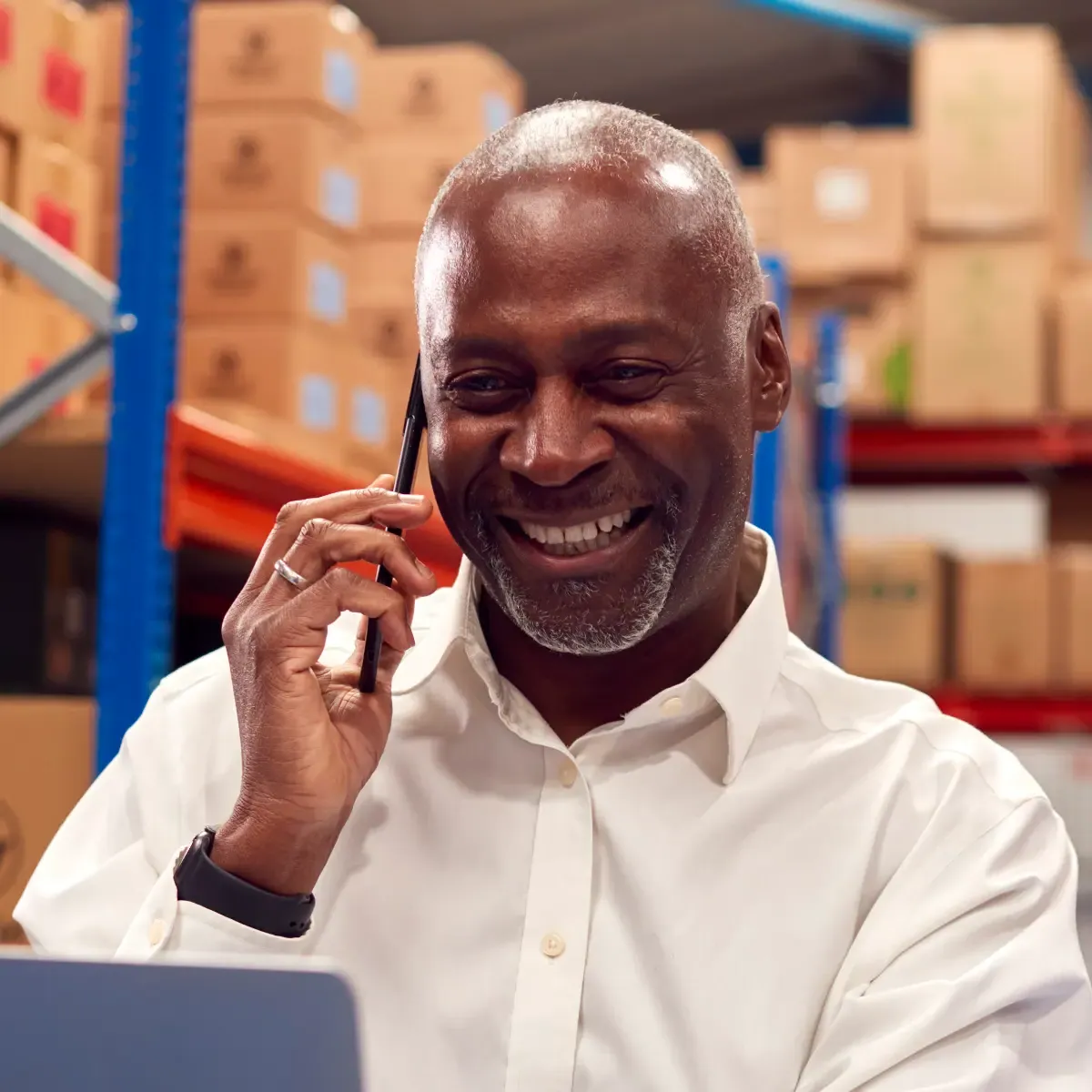 Older smiling man sitting at a desk inside a warehouse talking on phone and typing on a laptop.