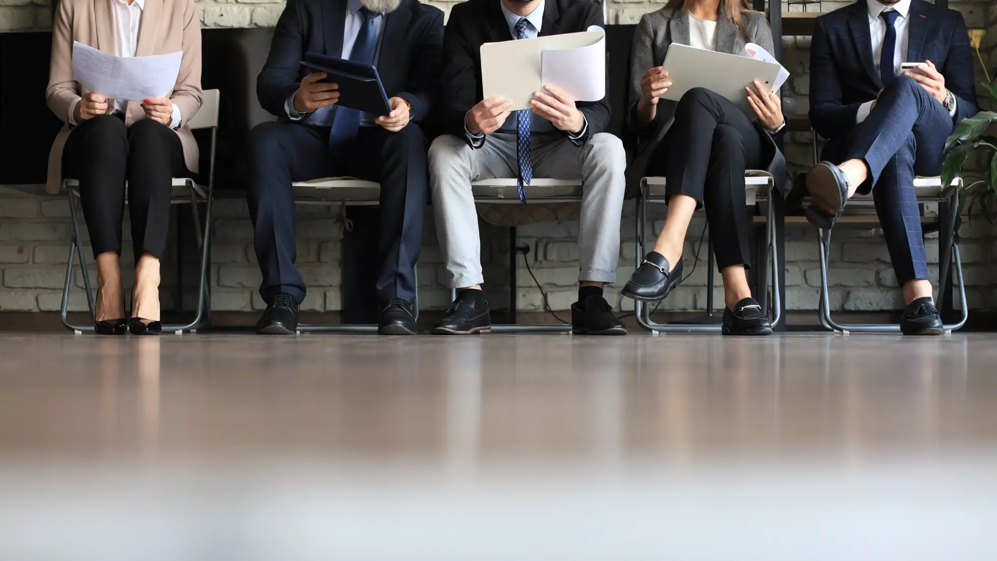 Bottom half of five applicants wearing professional outfits sitting in chairs waiting for job interviews.