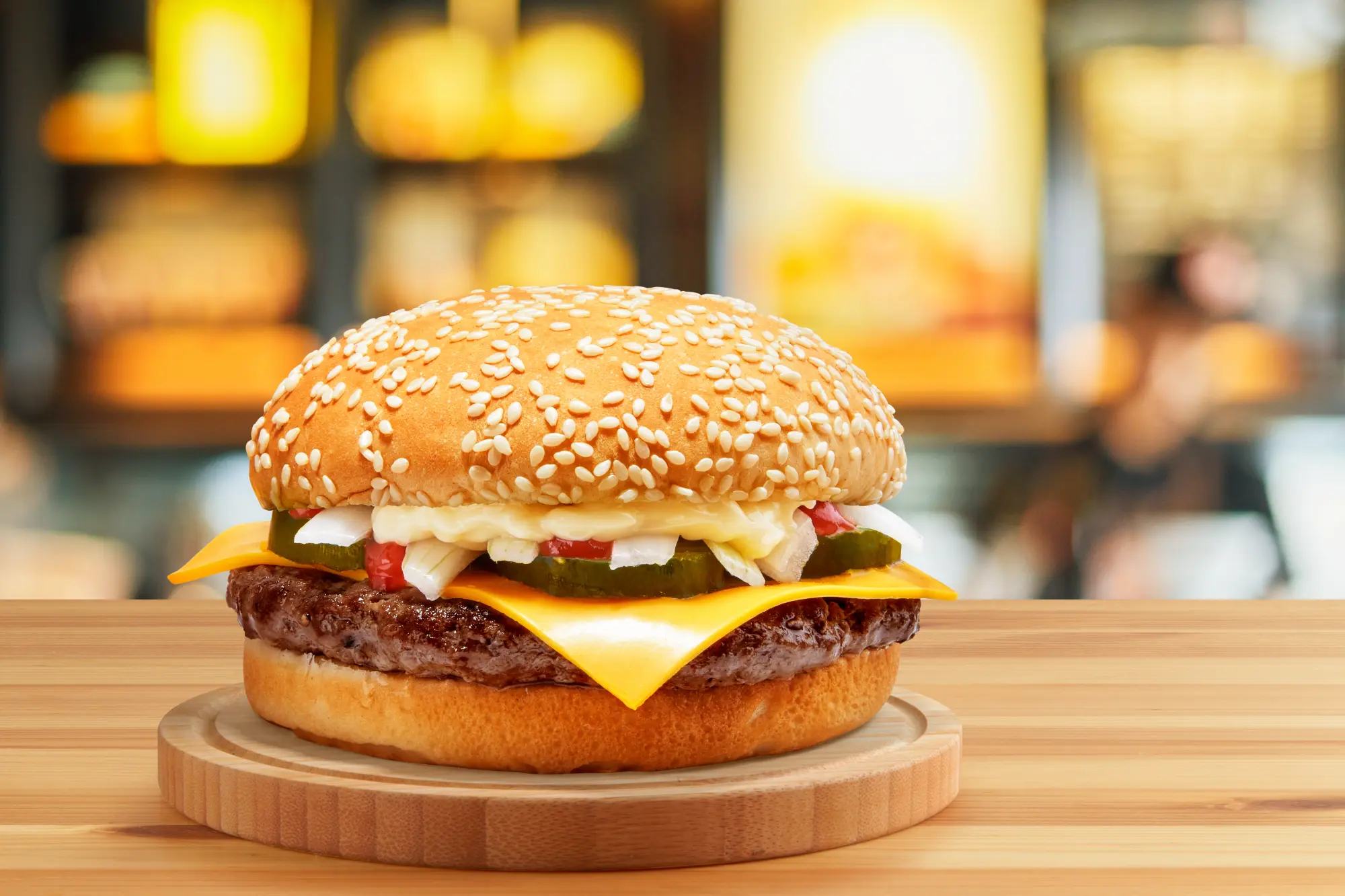 A large cheeseburger featuring a beef patty, cheddar cheese, pickles, diced white onions, and sauce, presented on a wooden board.